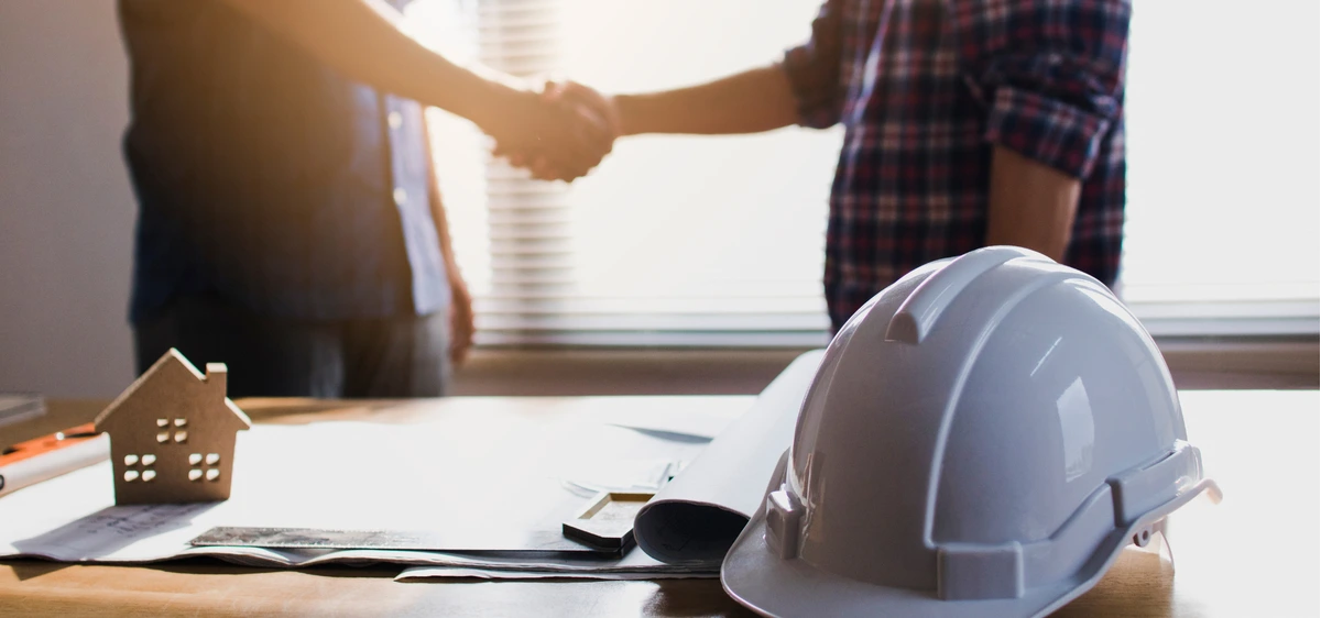 Handshake over blueprints, hard hat, and model house. Working with a licensed handyman.